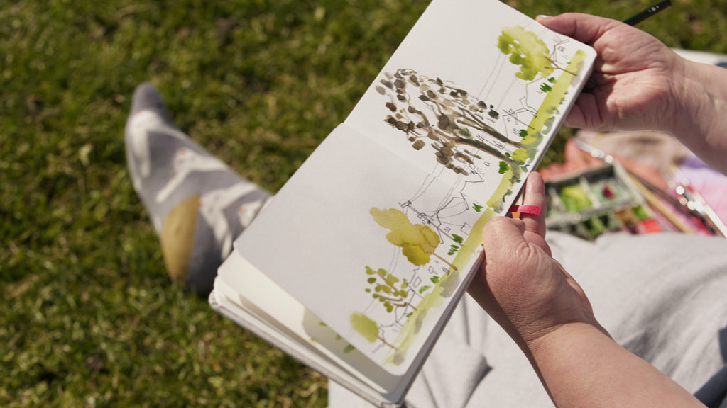 hands holding a sketchbook depicting a watercolor landscape