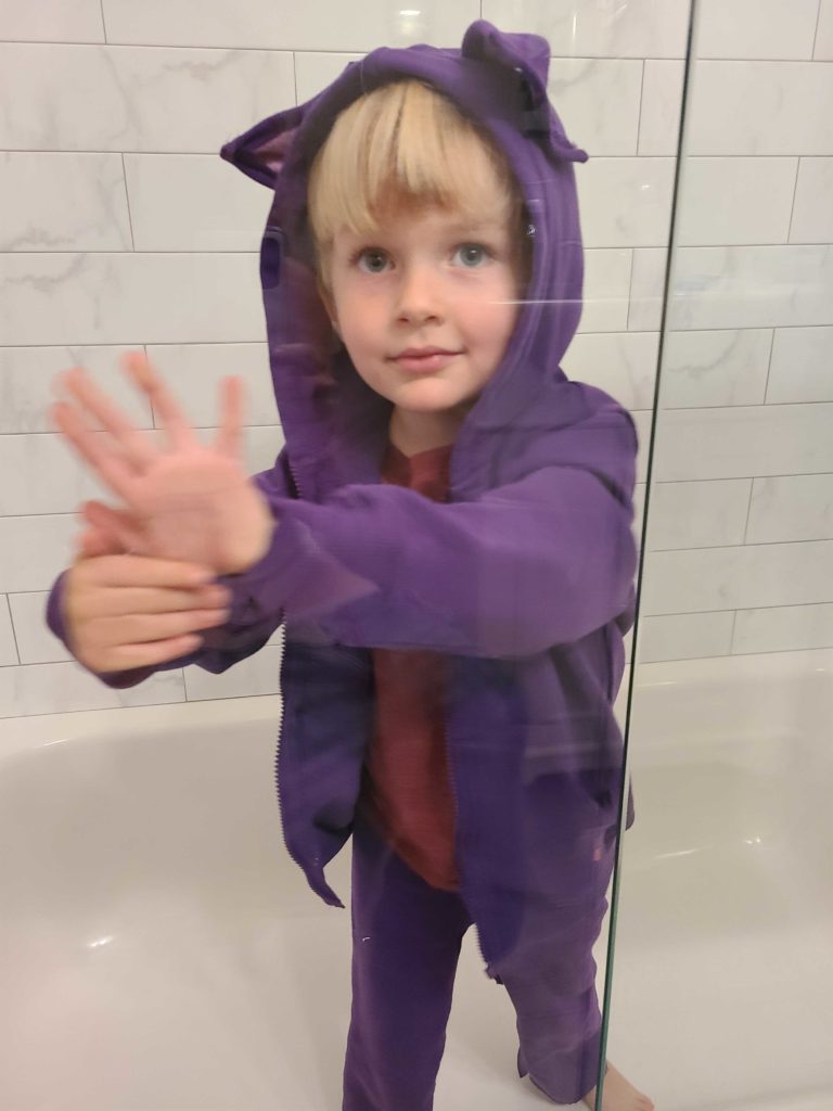 A young boy smiles while standing in a bathtub and wearing a homemade purple cat hoodie