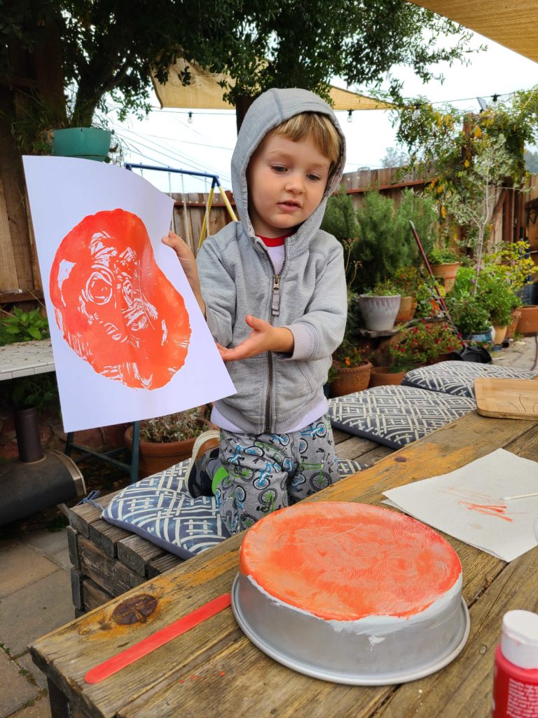 a child holding a mono print he made by painting orange onto the back of a pie tin