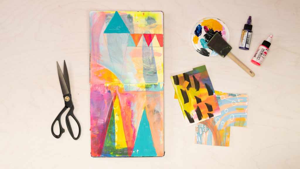An open sketchbook on a wooden table revealing colorful abstract art by Abby Houston alongside a pair of scissors, paint, and a sponge brush. 