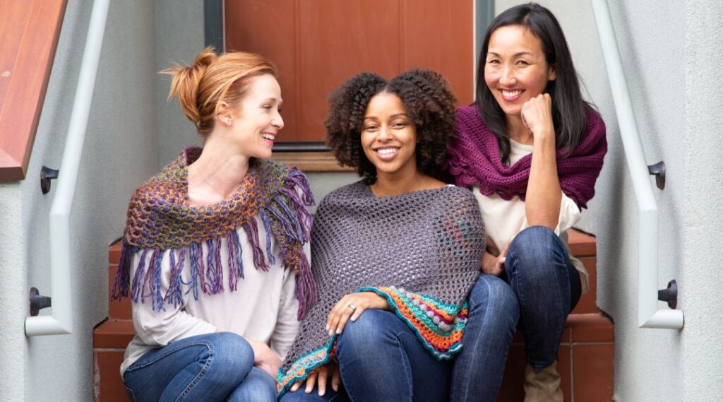 image of three women sitting on a stoop, smiling and wearing handmade crocheted shawls to illustrate how crocheting and knitting benefit your mental health
