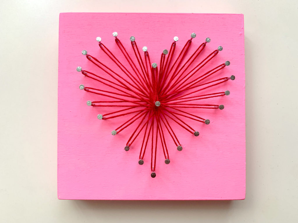 Heart-shaped string art with red thread and a pink wood board