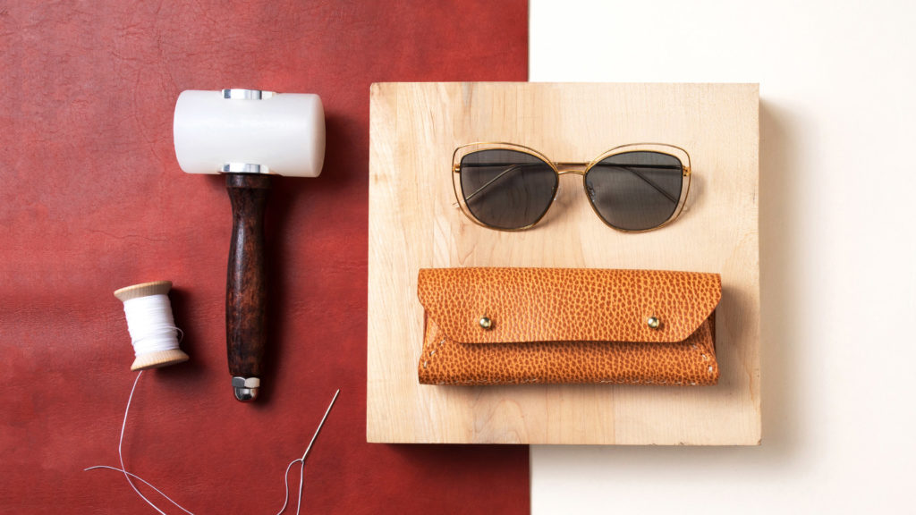 Flatlay including sunglasses, leather eyeglass case, mallet, wooden pound board, and thread