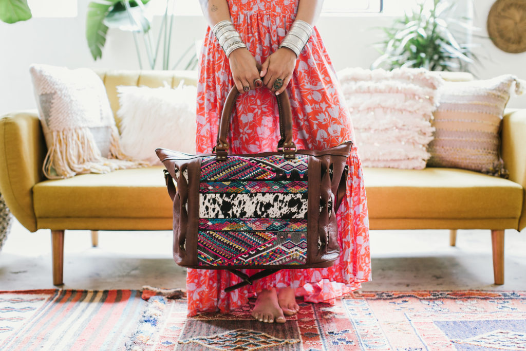 A bohemian-inspired leather bag with woven textile accents and hair-on hide, held by designer Treasure of Nikki&Mallory in front of a couch