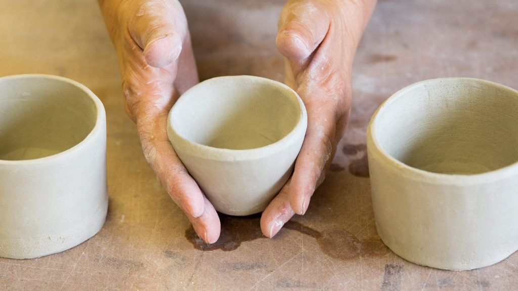 Close-up photo of a pair of hands holding a small cup made of clay, with 2 more hand-built ceramic cups or bowls on each side