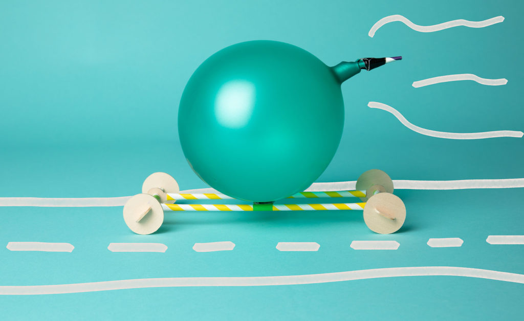 a teal balloon sits on top of a small, simple vehicle made of paper straws and wooden wheels on a teal background with white motion lines and a hand-drawn road
