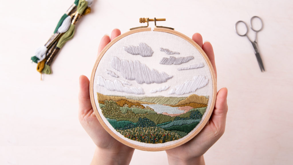 Flat lay on a white table of two hands holding a hand-embroidered landscape of green hills and grey clouds in an embroidery hoop