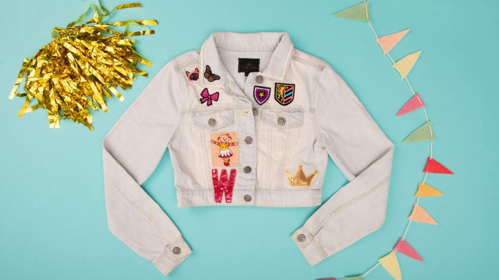 Flat lay on blue background of a denim jacket with hand sewn patches and trim plus a gold pom pom and rainbow garland