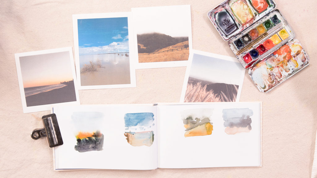 A flat lay of a sketchbook, landscape photos, and a watercolor palette on a neutral background