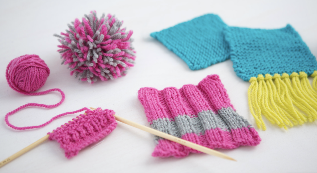 Flat lay on a white table showing how to knit with a small pink knit swatch on knitting needles, some small knit projects in blue and yellow, and a pink and grey pom pom