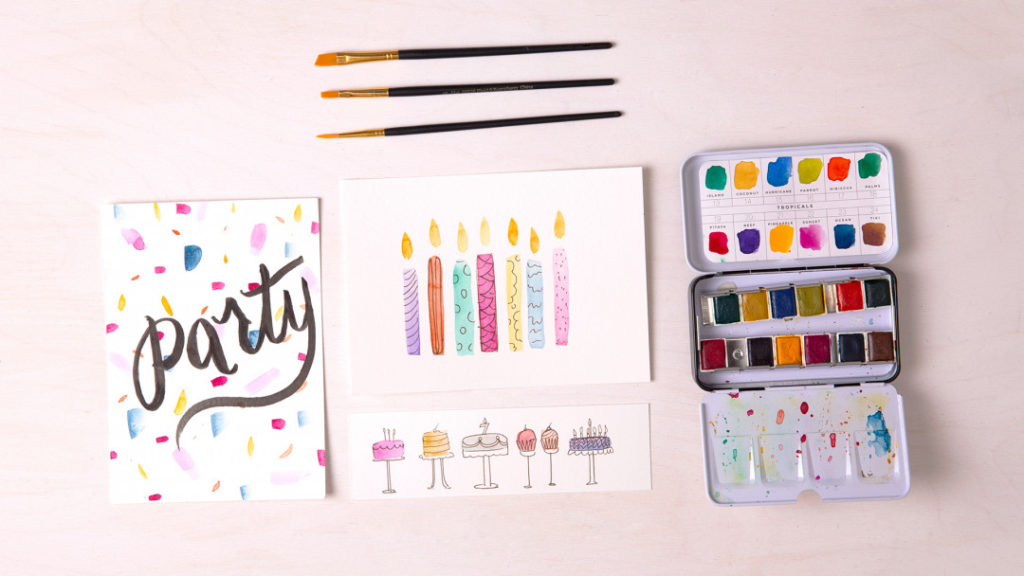 a flat lay of watercolor brushes, a small watercolor palette filled with paint, and a few hand painted birthday cards featuring candles, cake, and confetti