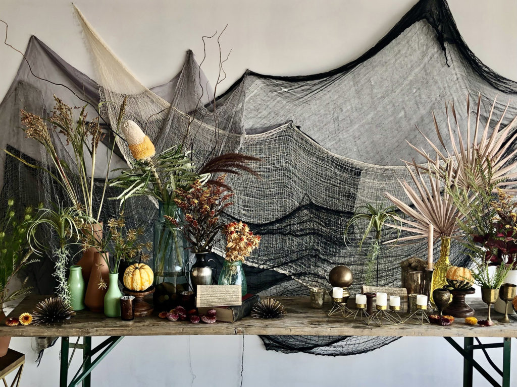 Backdrop for a film set with a spooky fall theme including black and cream gauze draped on the wall plus a wood table filled with various plants, gourds, and candles