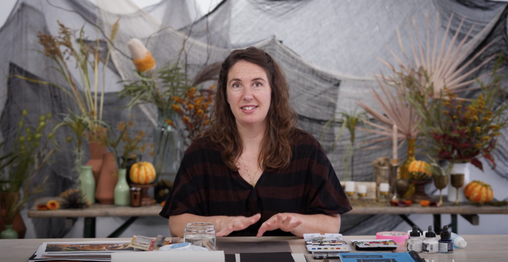 Courtney Cerruti sits at a table to teach a monoprinting class in front of a spooky backdrop with gauze, plants, candles and gourds