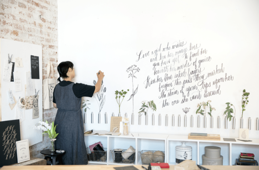 A woman stands in front of wall with calligraphy on it while drawing flowers in black ink