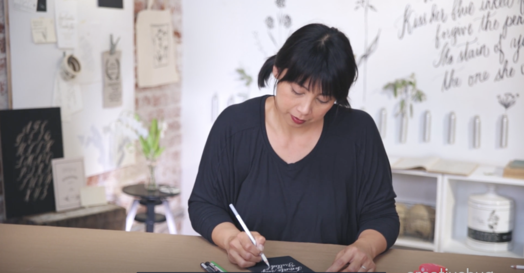 Maybelle Imasa-Stukuls in a dark shirt working on a calligraphy project in front of a white wall with her art pinned up