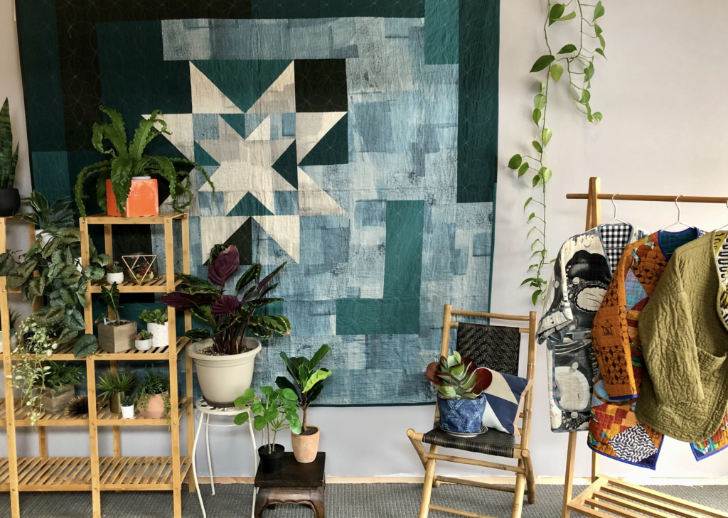 A set for a film shoot that includes a large blue-toned patchwork quilt wall hanging, bamboo shelf filled with plants, and a rack on the right with hanging coats and jackets