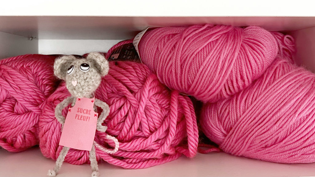 A crocheted mouse wearing a pink baking apron stands in front of pink balls of yarn.