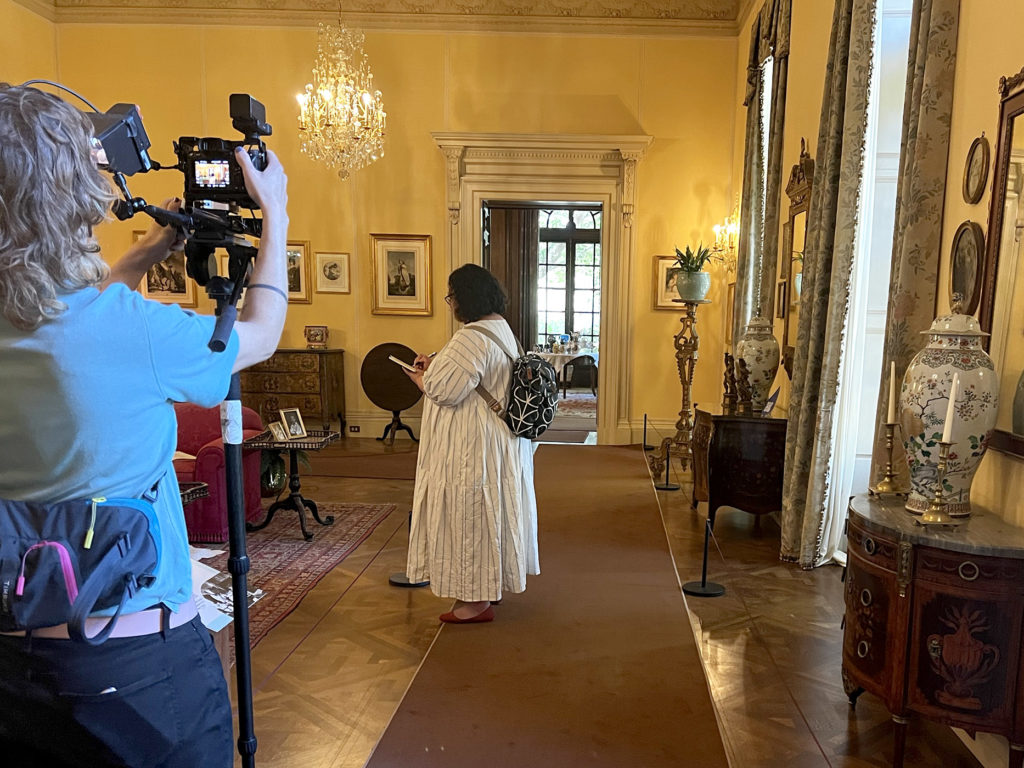 Mariko Jesse stands and sketches inside the ornate room of a mansion while Sam films her with a video camera.