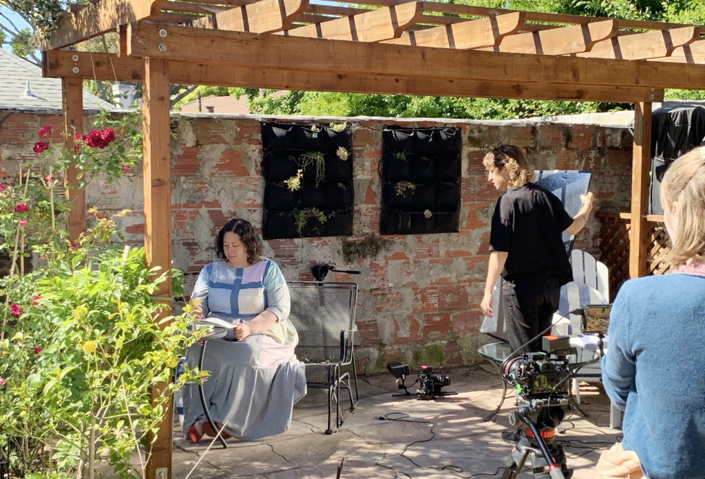 Three people in a sunny garden underneath a wooden pergola next to a brick wall