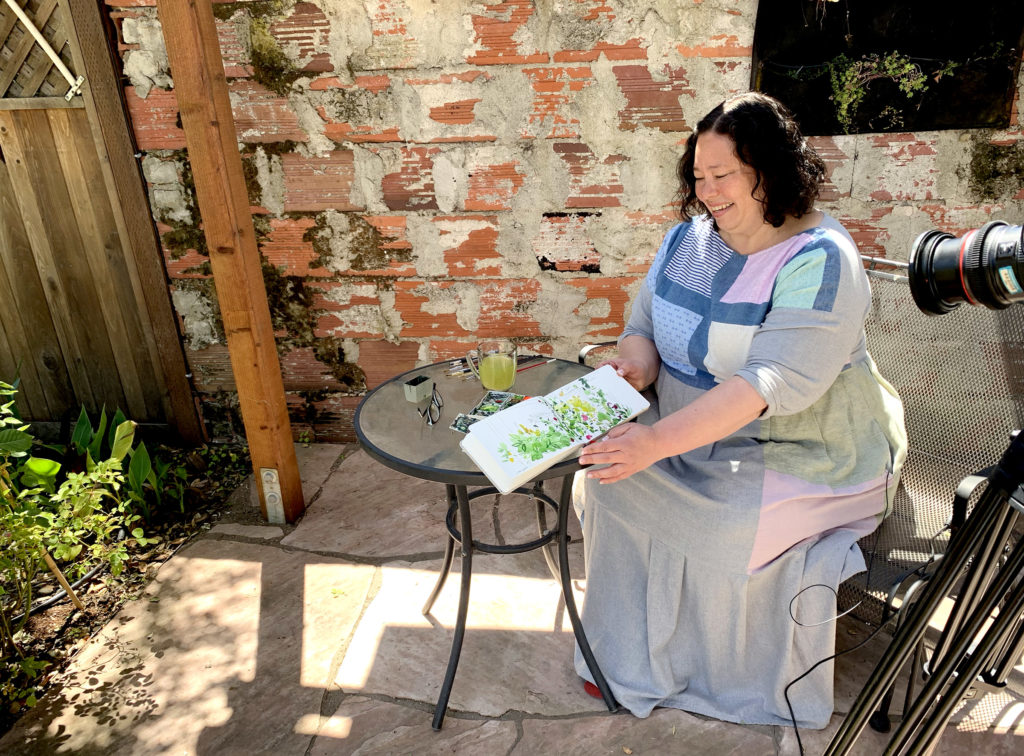 Artist Mariko Jesse smiles and sits at a garden table, showing her colorful watercolor sketch of plants and flowers