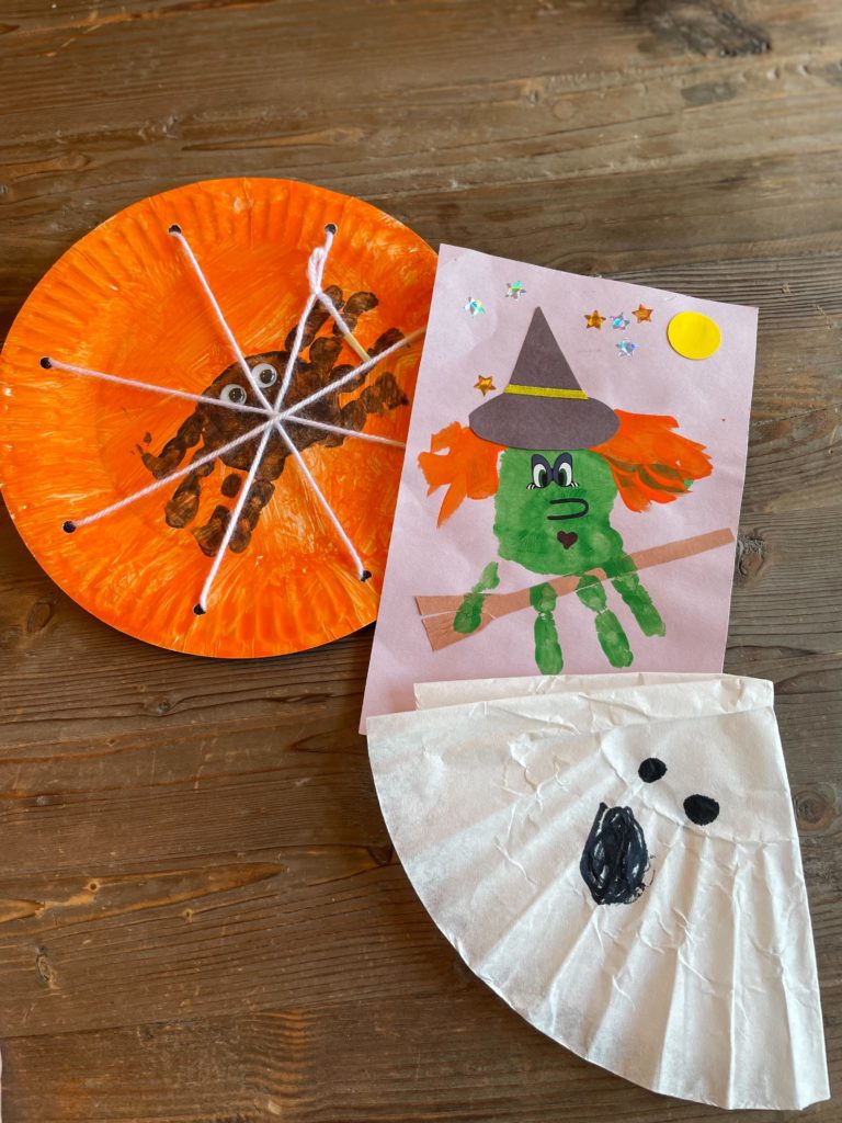 Halloween kids crafts on a wooden table including a green handprint witch, a coffee filter ghost, and a painted spider on a paper plate