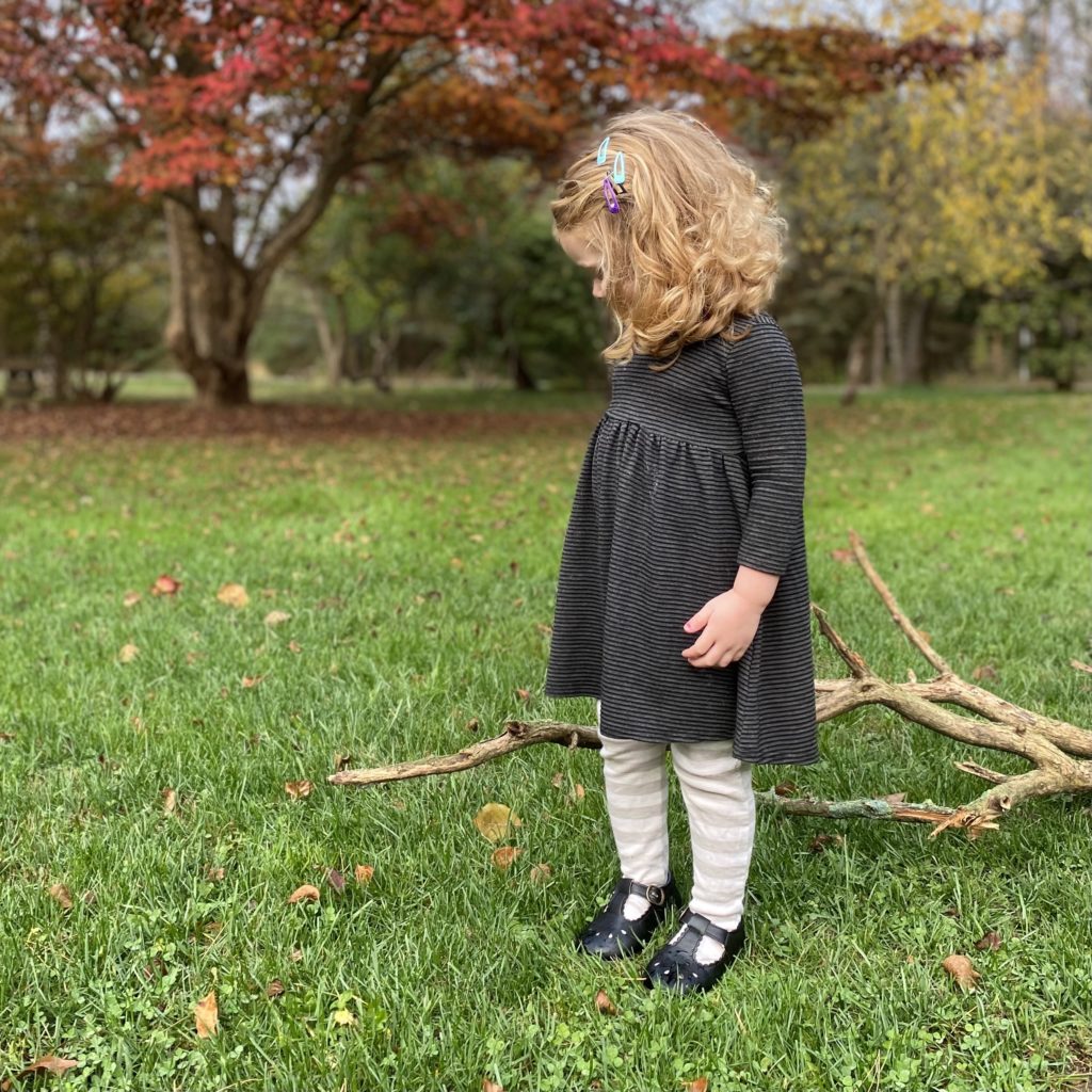 small child with curly blonde hair in an autumn grassy field wearing a dark grey striped tunic that was sewn from an adult sized tunic