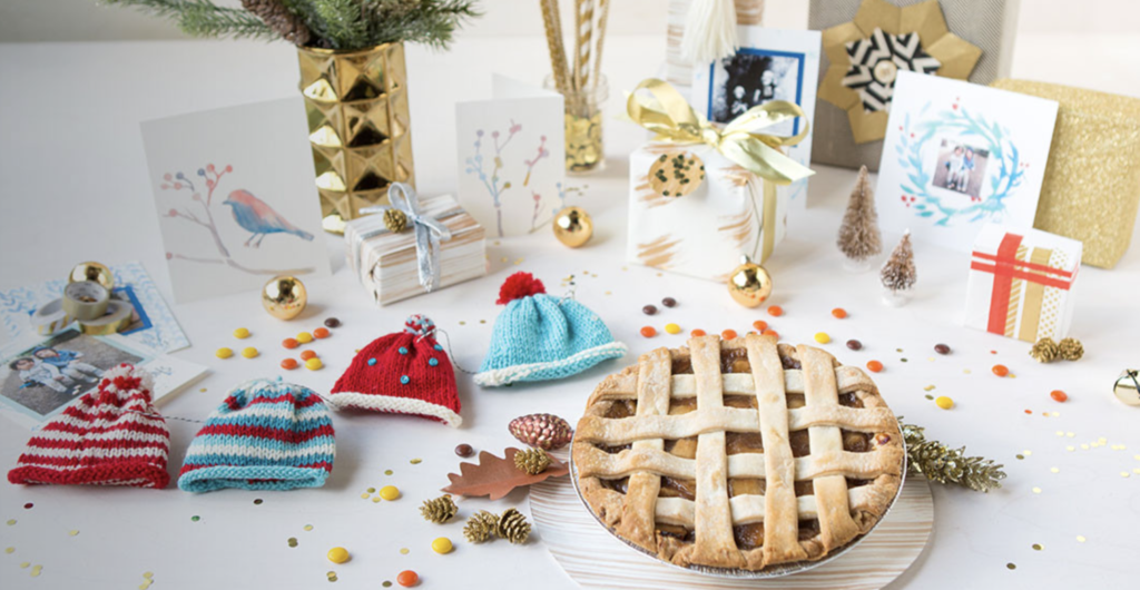 a holiday display of handmade goods like a knitted mini hat garland, and apple pie, watercolor holiday cards, confetti, and gold and silver embellishments