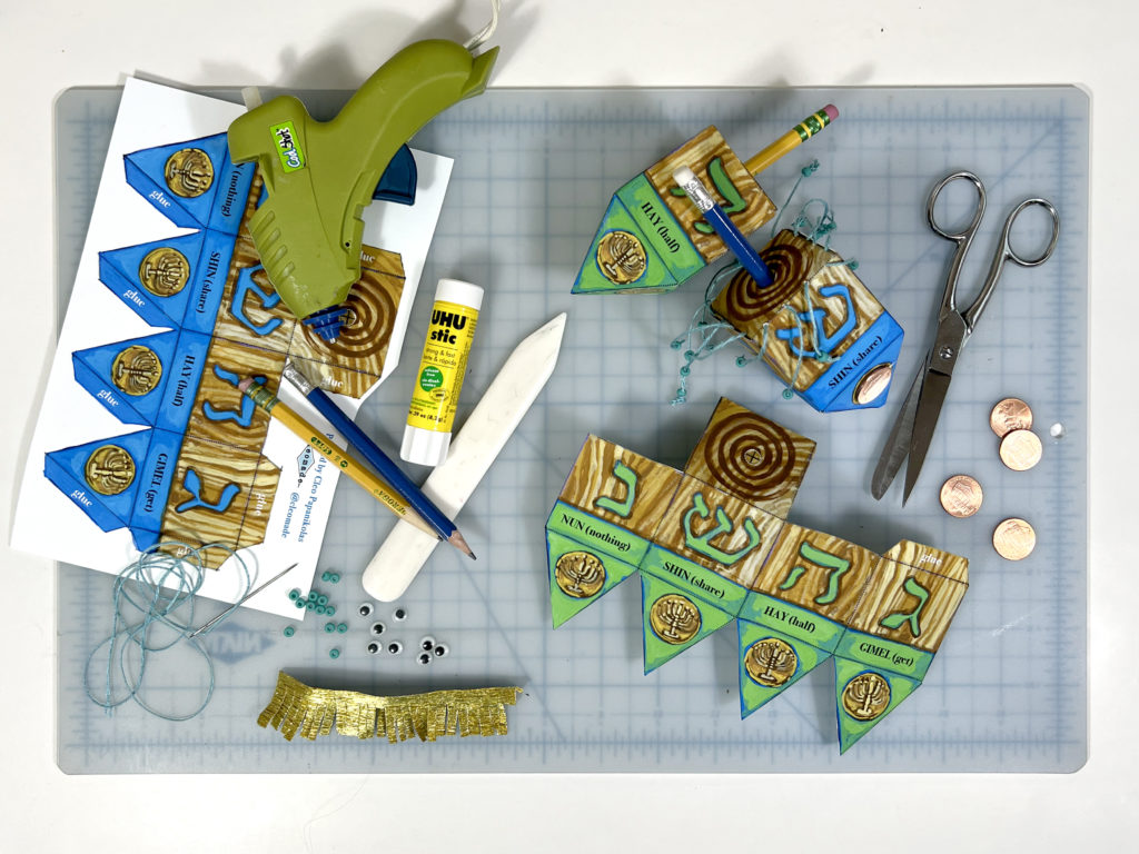 flat lay of supplies on a gridded cutting mat for printing and making your own paper dreidel such as glue stick, glue gun, scissors, crepe paper fringe, googly eyes, and pennes