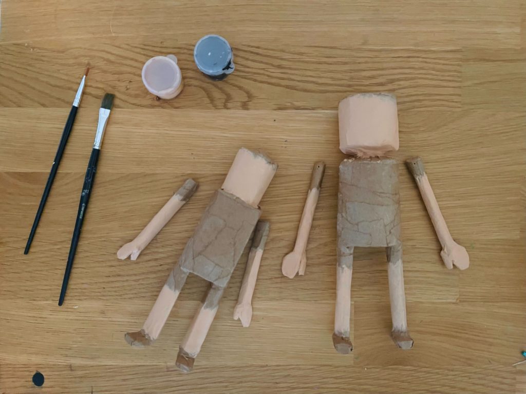 2 partially finished paper mache dolls on a wooden table with paint pots and paint brushes.