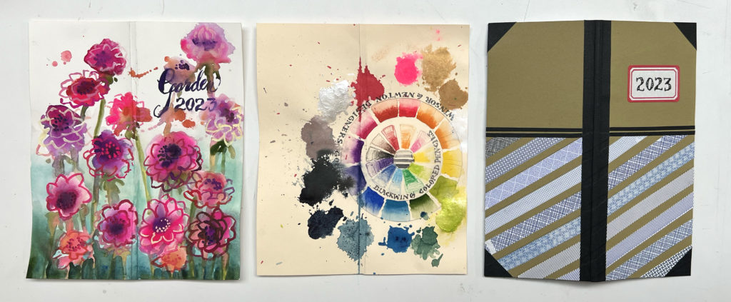 three handmade calendar/planners with different covers lying flat: on the left, pink and purple painted flowers, in the center, a hand-painted color wheel and paint splotches, on the right, a collage including strips of security envelopes on an olive green background and a label that says 2023.