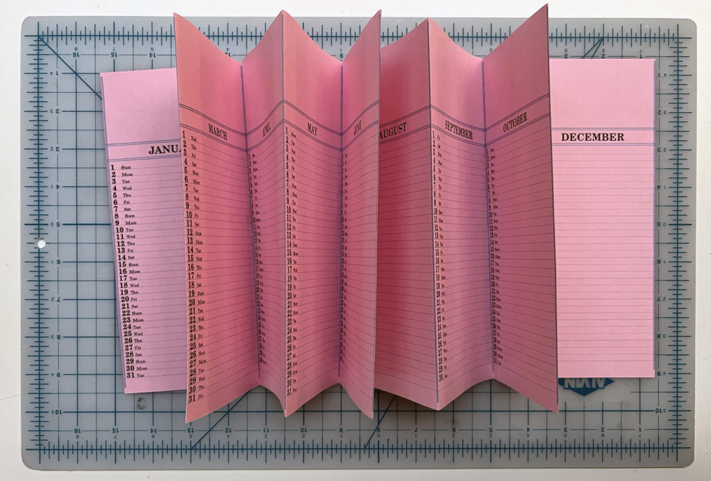 pink printed calendar pages formed into an accordion book, lying on top of a gridded cutting mat