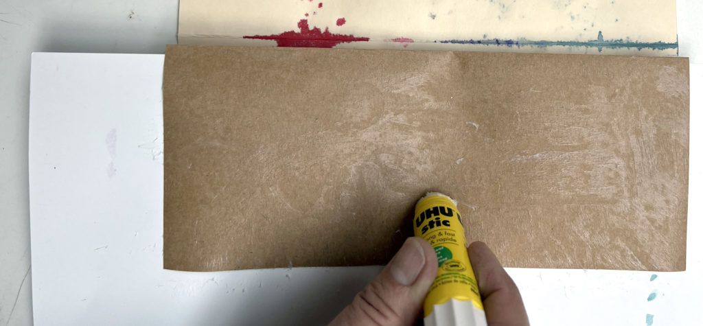 a hand holding a glue stick while gluing a brown piece of rectangular paper