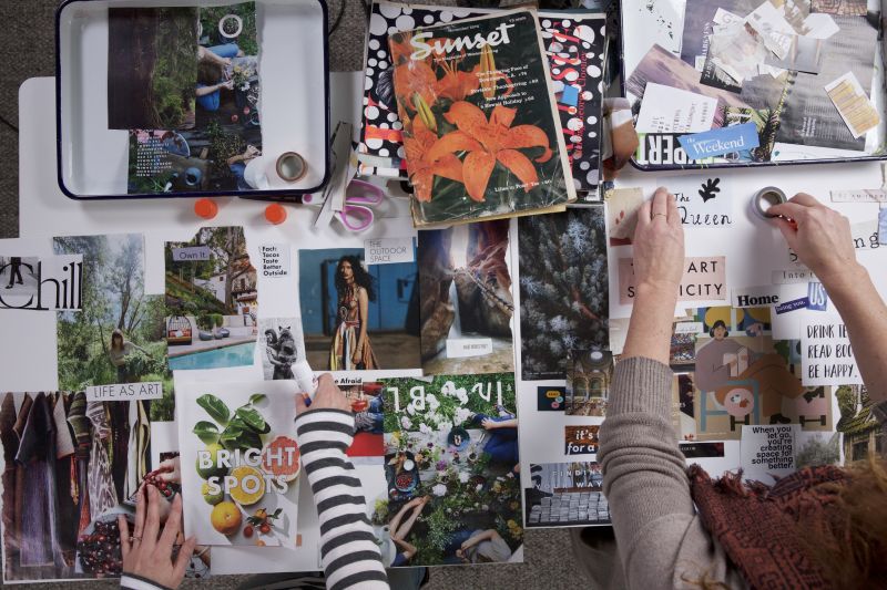 Flat lay featuring lots of magazine pages spread across a surface to create vision boards for 2023 along with the arms of two people arranging items. 