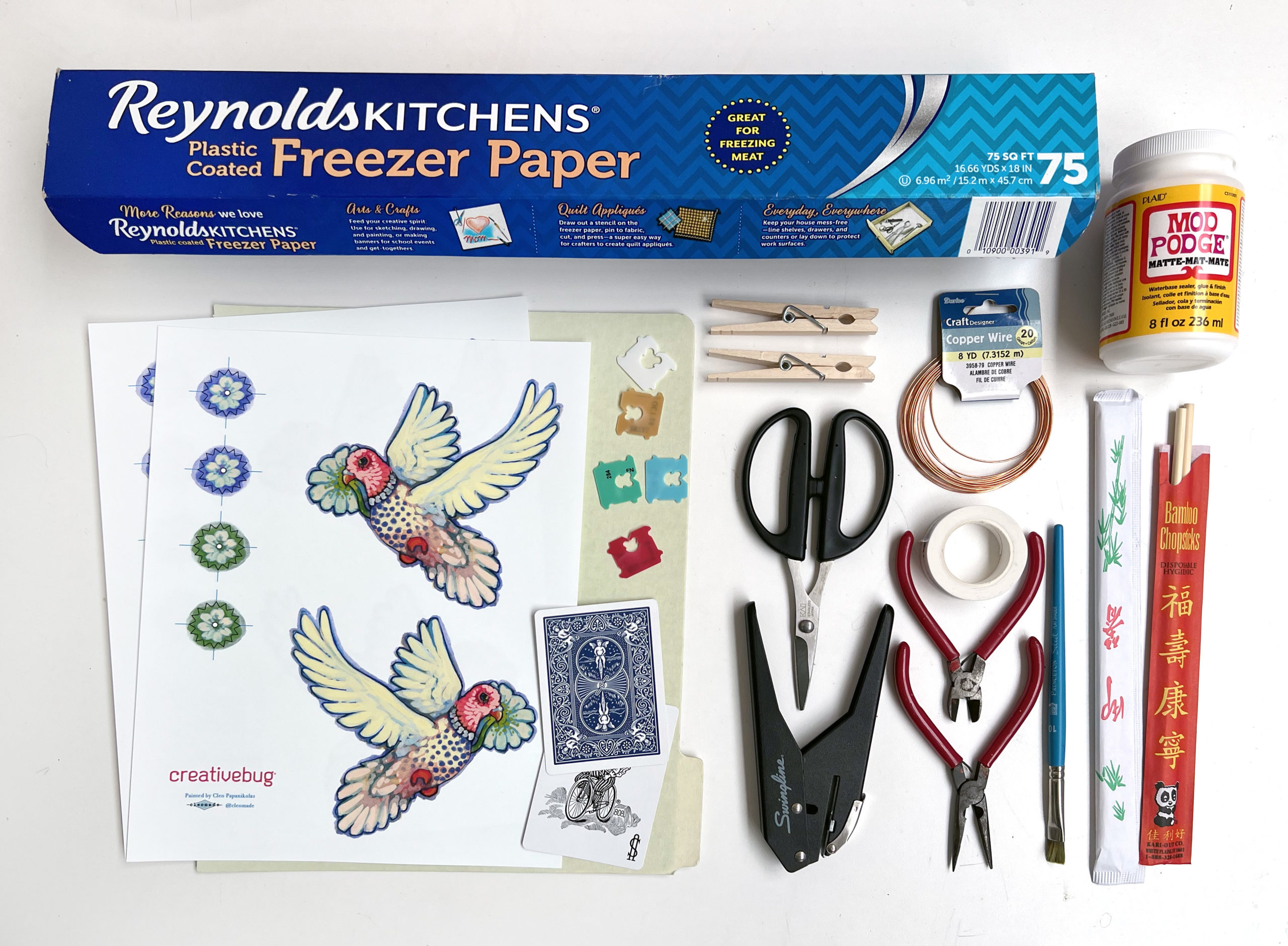 a flat lay of materials to make a noisemaker or grogger for Purim including a free paper template, freezer paper, cardstock, mod podge, clothespins, wire cutters, pliers, chopsticks, wire, and a hole punch