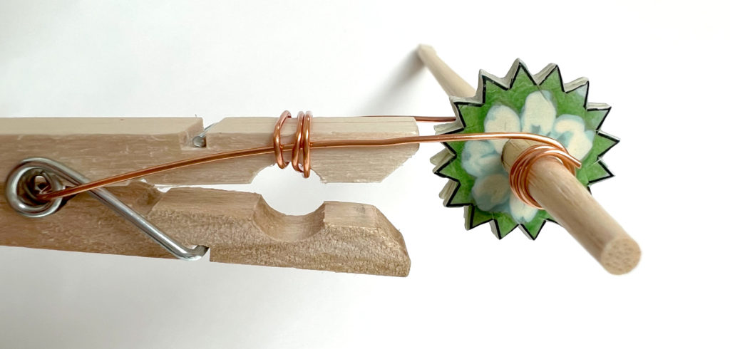 close-up of a noisemaker mechanism with a wooden chopstick, wire, clothespin, and DIY paper gears