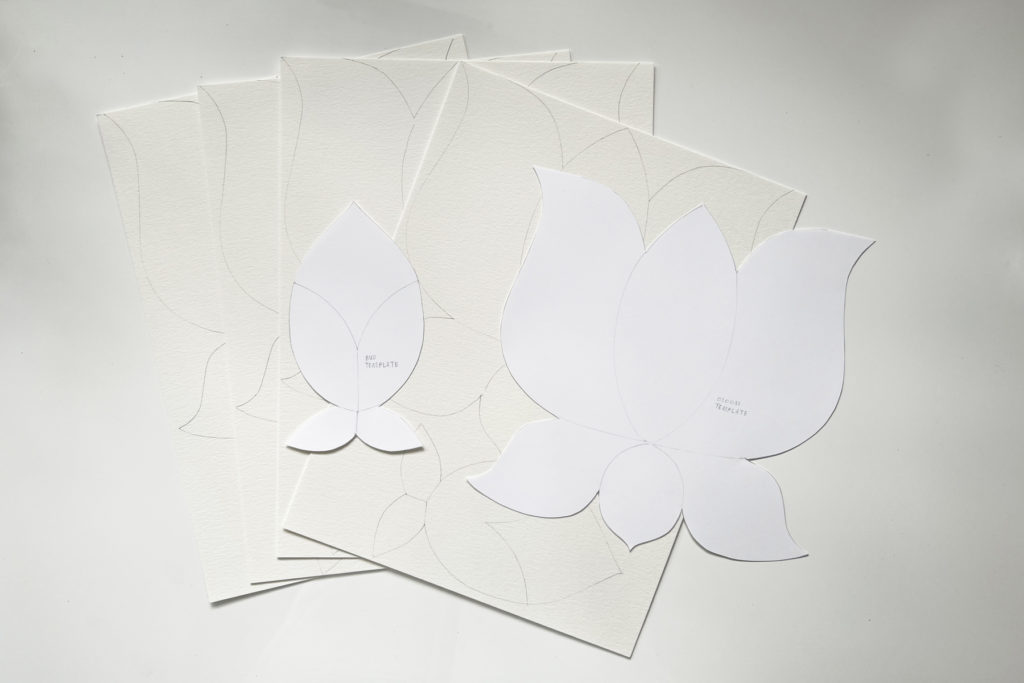 flat lay on a white table of white paper templates for a lotus flower and lotus bud on top of a stack of 4 traced flowers on watercolor paper