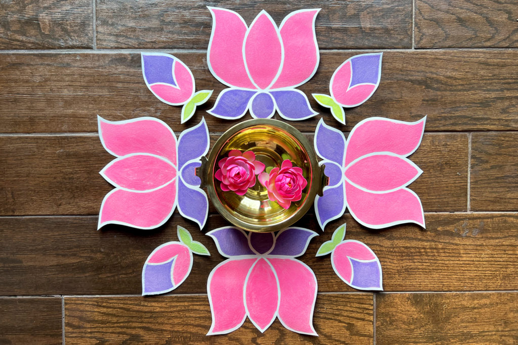 lotus-shaped sand art rangoli on a dark wooden floor in a square design with a brass bowl of water and pink flowers in the center