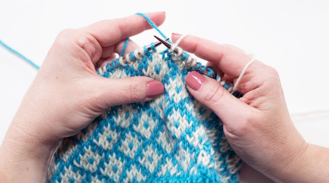 image of instructor Edie Eckman's hands knitting a fair isle project in blue and cream illustrating how occupying your mind with crocheting or knitting has many mental health benefits