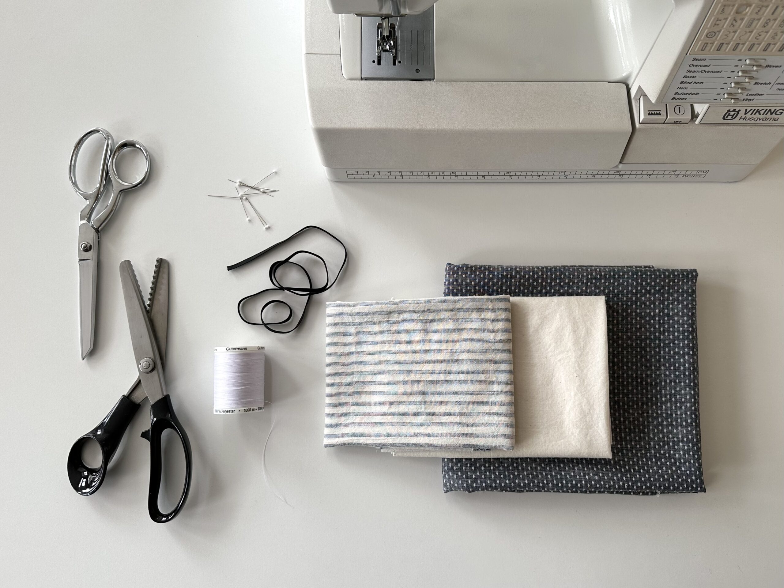 image flatlay of sewing machine, scissors, fabric shers, pins, elastic, thread, and fabric in grey stripe and navy polka dots to sew doll clothing