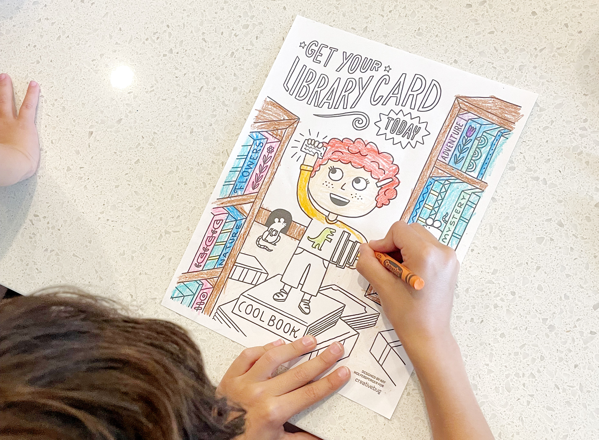 image of a child holding an orange crayon and coloring a page depicting a cartoon child holding a library card between two bookshelves
