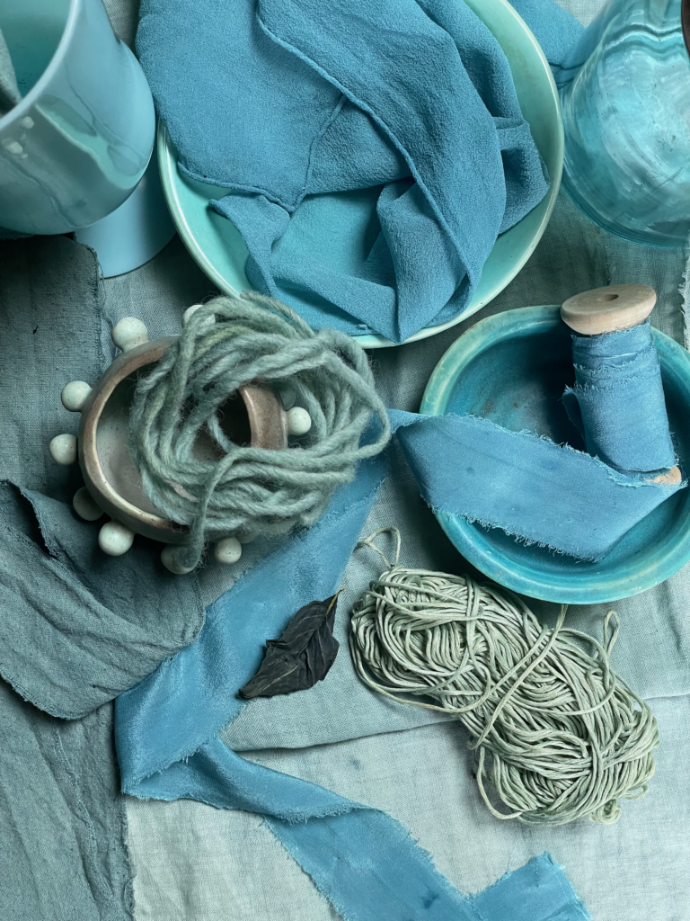 image of various textiles dyed teal blue with fresh indigo leaves: yarn, strips of fabric