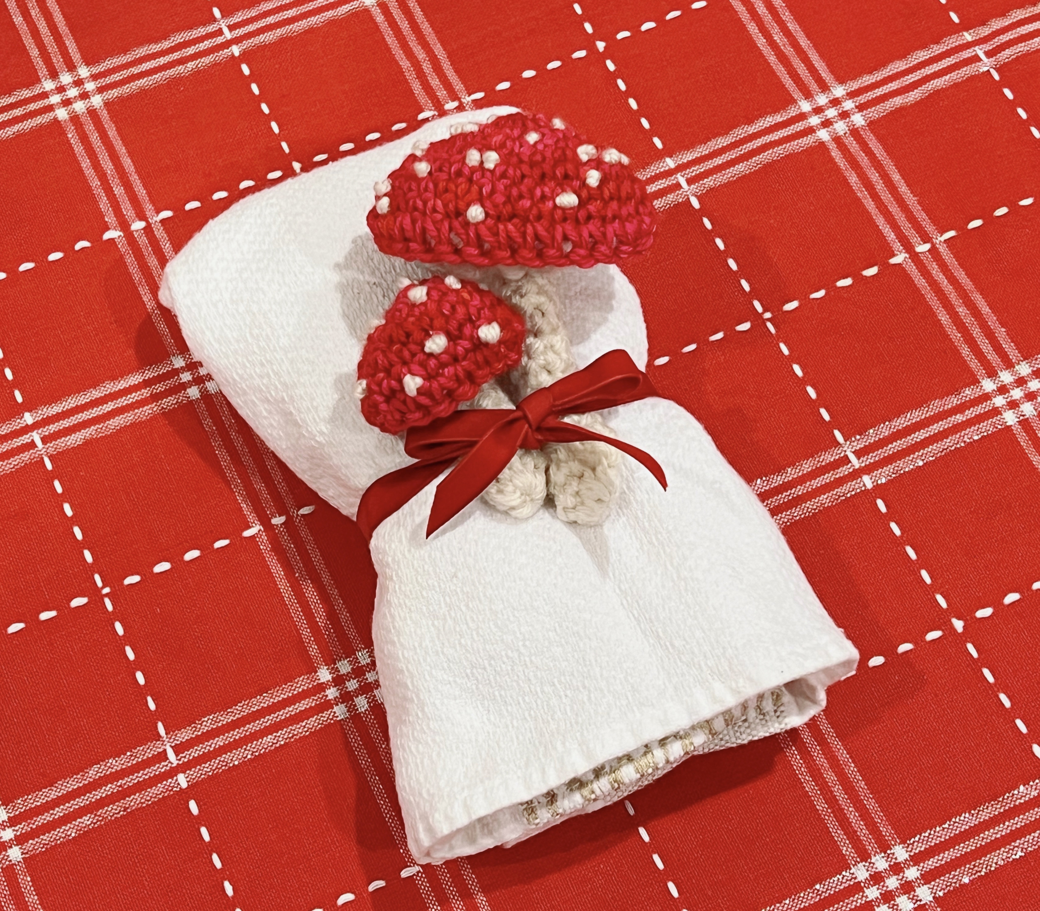 image of two small crocheted toadstools tucked under a red velvet ribbon around a white napkin on a red and white gingham tablecloth