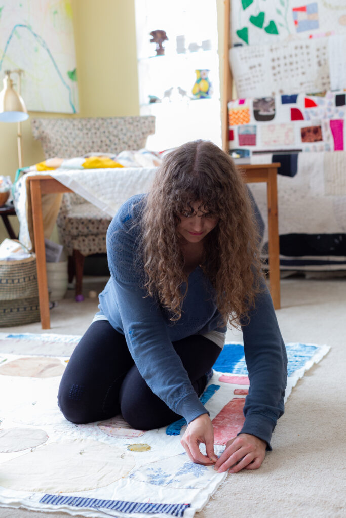 a woman in a sunlit craft room kneeling on the floor on a quilt she made