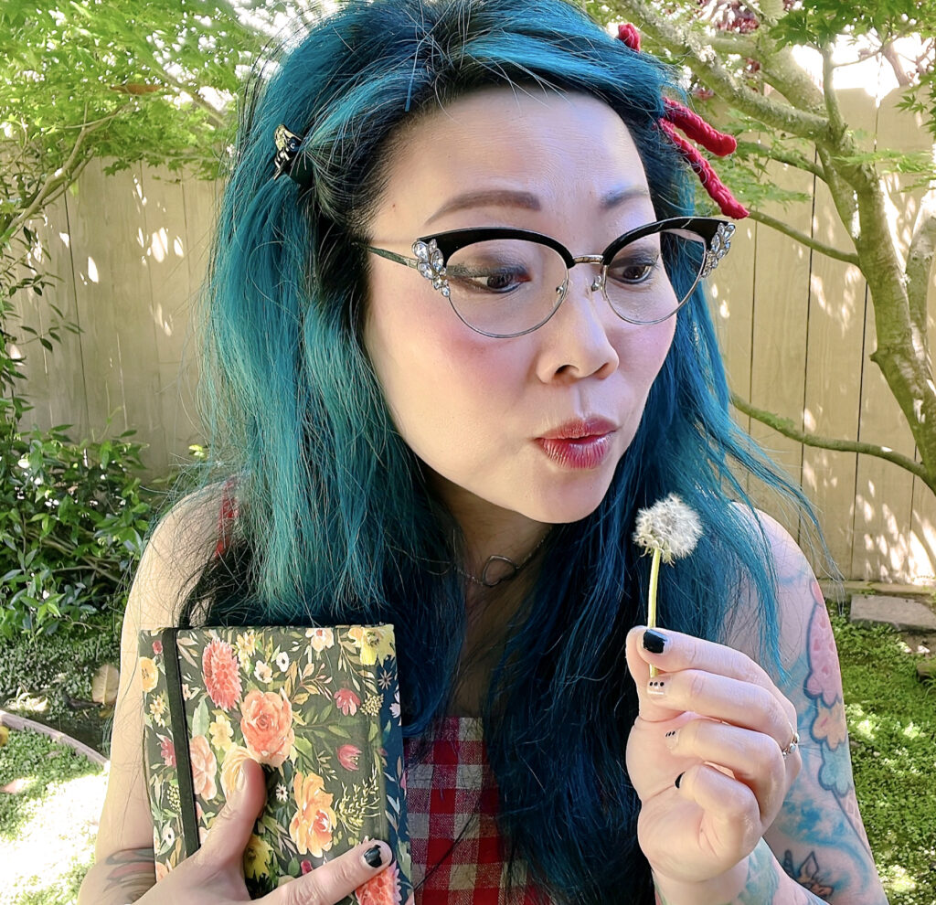 image of a person with blue hair and glasses, blowing on a dandelion while holding a floral travel journal