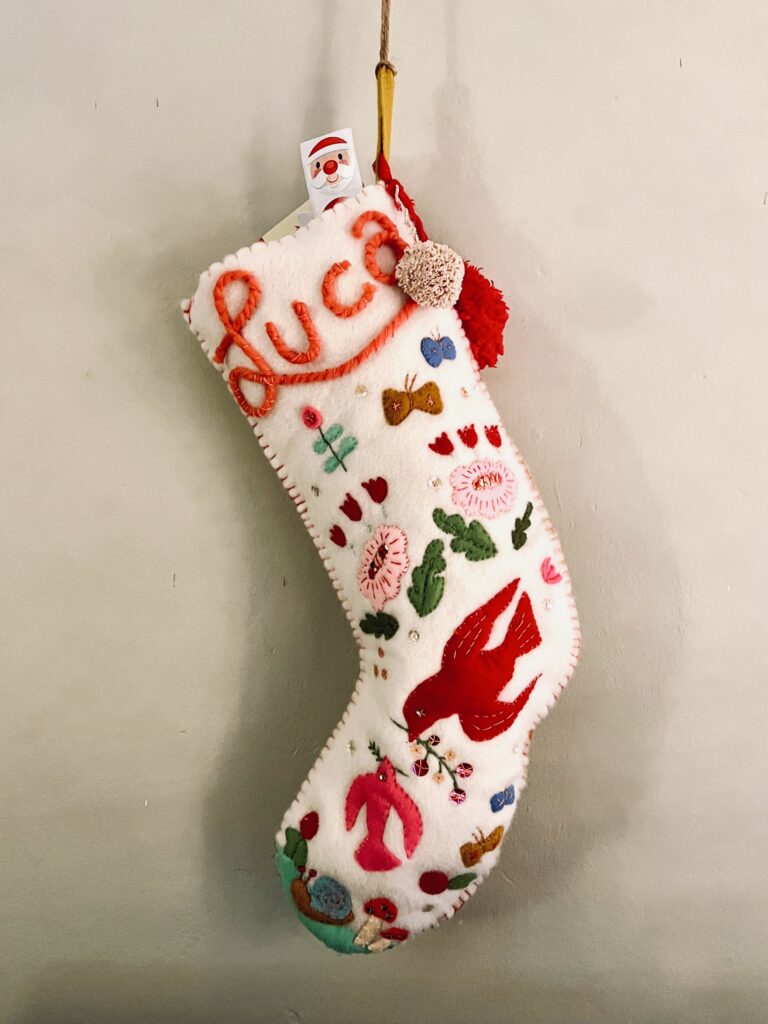 image of finished handmade heirloom stocking in off-white felt covered in many colorful felt appliques like birds, flowers, and snails, hanging against a wall