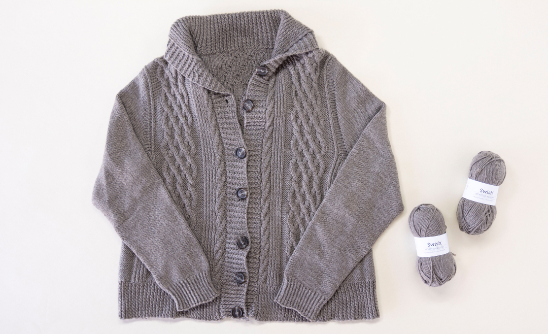 image of a grey, hand knit cardigant with a cabled texture on a white table top