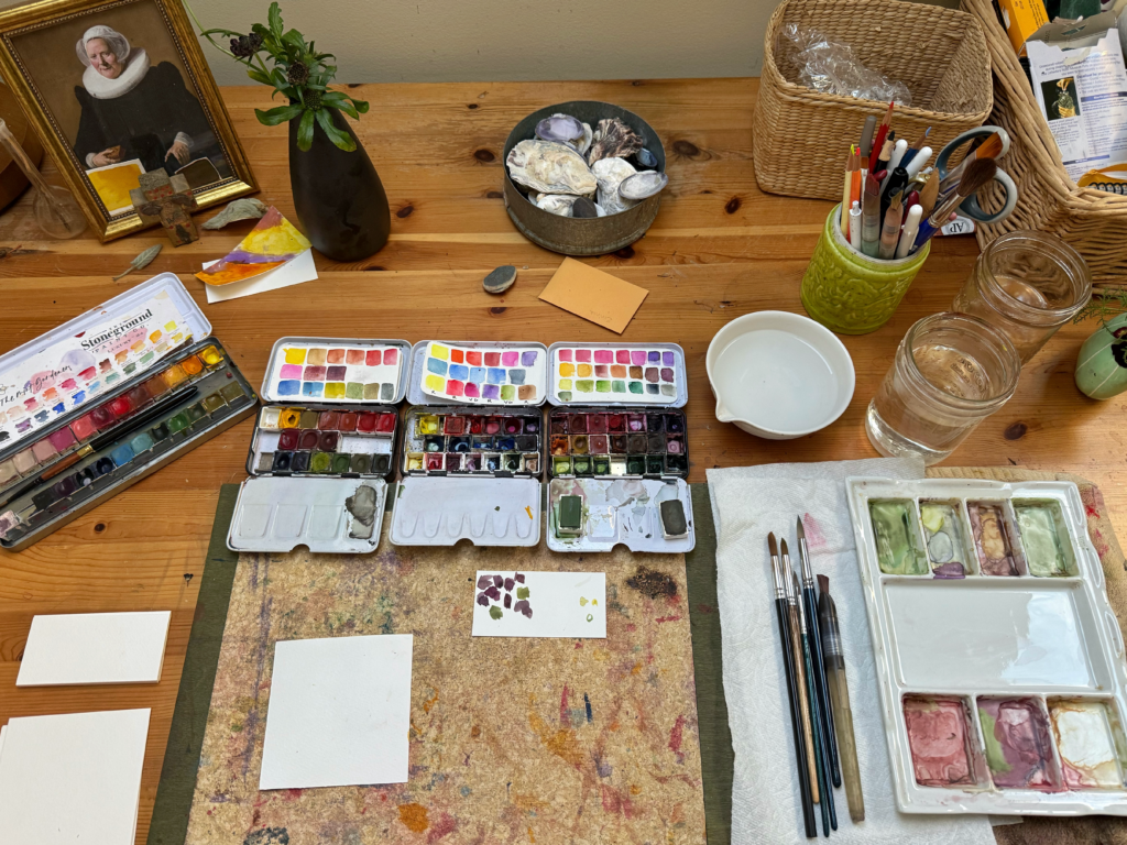image of a wooden work table with many watercolor palettes and brushes, ready for a daily color study session