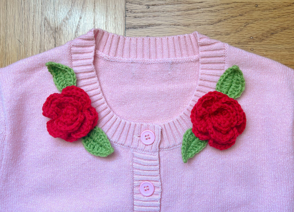close-up image of a pink knit cardigan decorated with 2 large red crocheted roses and green leaves